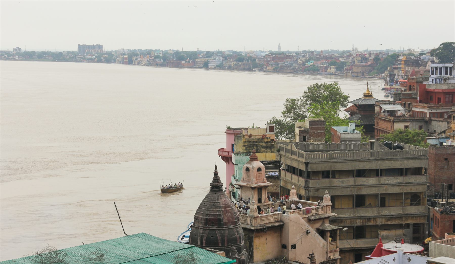 Riverside buildings and temples of Varanasi above the wide Ganges, with a small boat on the water