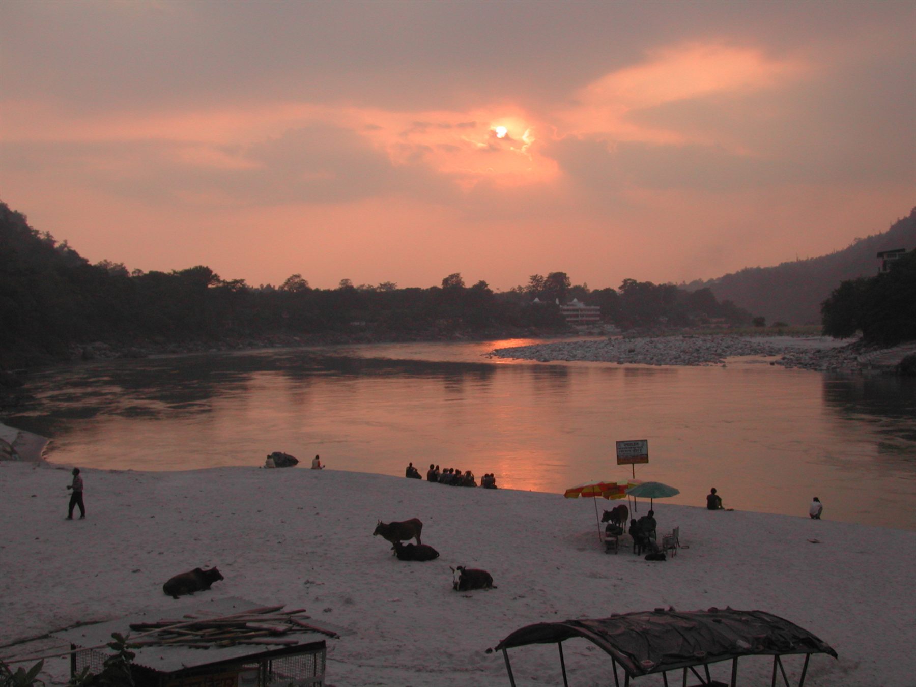 Sunset over the river between wooded hills, with cattle and people on the sandy shore